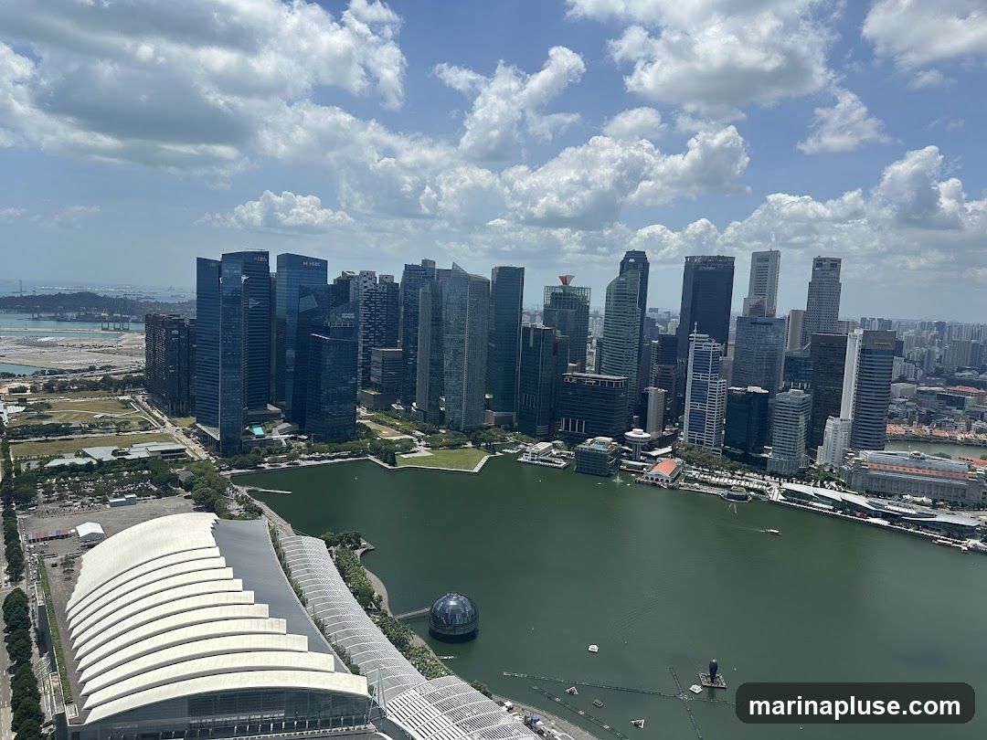 An aerial view of a modern cityscape with numerous skyscrapers and a large body of water. In the foreground, a large, curved white structure with a ribbed roof dominates the lower left of the frame. Beyond this, a green body of water, likely a bay or river, stretches across the middle ground. Across the water, a dense cluster of tall, modern buildings forms the city skyline. Some of these buildings have visible signage, including 'UOB' and 'HSBC'. The sky above is blue with scattered white, fluffy clouds. In the distance, more buildings and a hint of land can be seen.