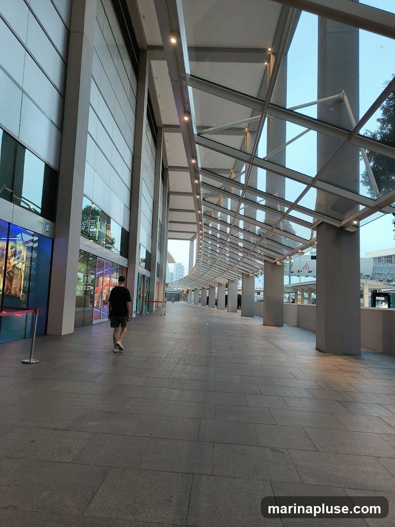 A person walks down a wide, paved walkway in front of a modern building with a glass and metal facade. The walkway is covered by a sloped glass roof supported by pillars. The building features large windows and a sleek, contemporary design. The sky is visible through the glass roof, indicating it is daytime.