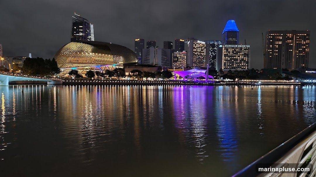 A nighttime view of a city skyline across a body of water. The prominent structures include a domed building with a golden facade, several modern skyscrapers, and a building with a distinctive blue illuminated top. The buildings are lit up, and their reflections shimmer on the dark water. A bridge is visible on the left, and a purple illuminated structure is in the mid-ground. The overall scene is illuminated by artificial lights, creating a vibrant and reflective urban landscape.