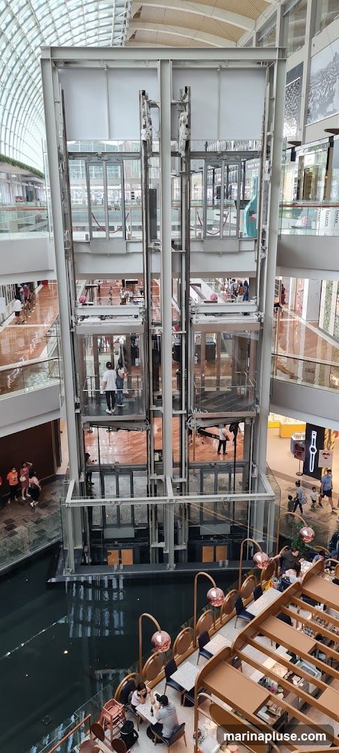The image shows the interior of a multi-level shopping mall. A large, glass-enclosed elevator shaft with multiple elevators is the central focus. People are seen inside the elevators and on the various levels of the mall. Below the elevators, there is a water feature. In the foreground, a cafe area with tables and chairs is visible, with patrons seated and enjoying their time. The mall has a modern architectural design with a glass ceiling allowing natural light to filter in.