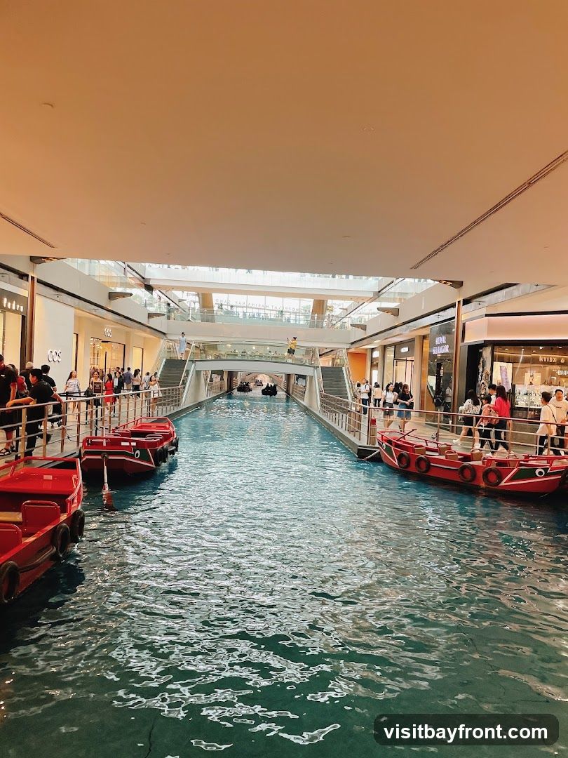 An indoor canal with red boats sailing on the water. People are walking along the elevated walkways on either side of the canal, which are lined with shops. The architecture suggests a modern shopping mall with a unique water feature.