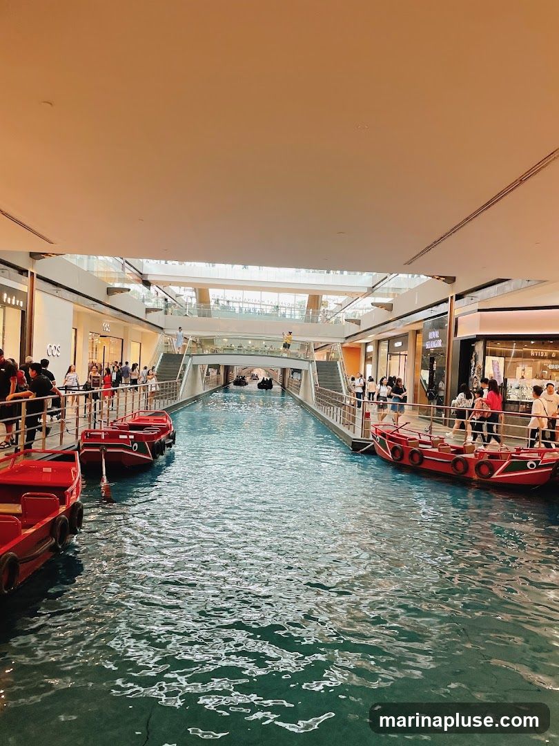 An indoor canal with red boats sailing on the water. People are walking along the elevated walkways on either side of the canal, which are lined with shops. The architecture suggests a modern shopping mall with a unique water feature.