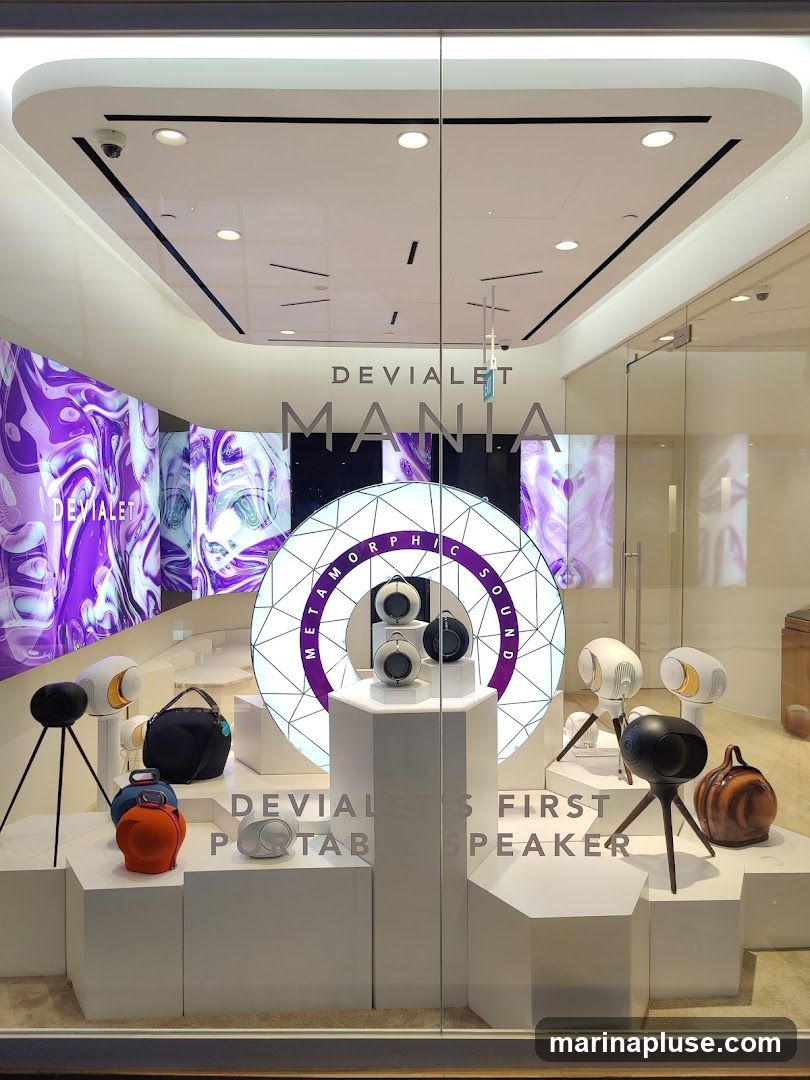 A retail display window showcasing Devialet Mania portable speakers. The display features several speakers in various colors and designs, some mounted on tripods. A large circular graphic with the text "METAMORPHIC SOUND" is prominent. The text "DEVIALET MANIA" and "DEVIALET'S FIRST PORTABLE SPEAKER" are visible on the window. The background includes abstract purple and white digital art displays.