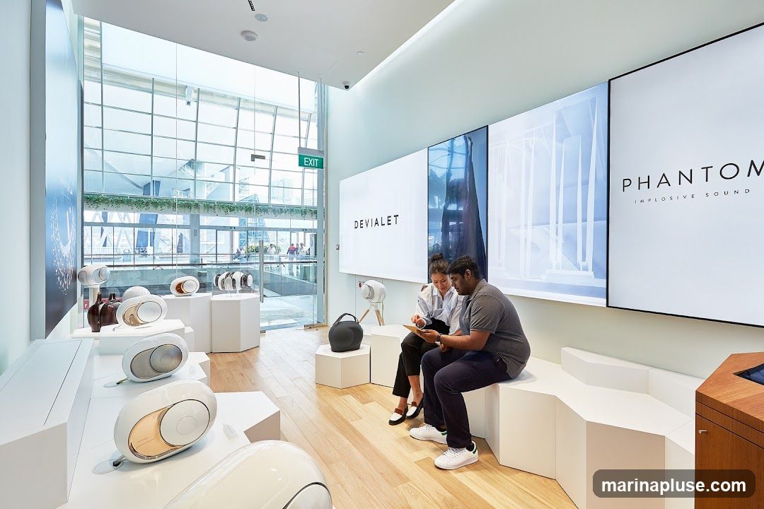 A modern, minimalist store interior with white hexagonal display stands showcasing various white and gold speakers. Two people are seated on a white bench, looking at a tablet. Large screens on the wall display the brand name "DEVIALET" and "PHANTOM IMPLOSIVE SOUND". An "EXIT" sign is visible through the glass facade.