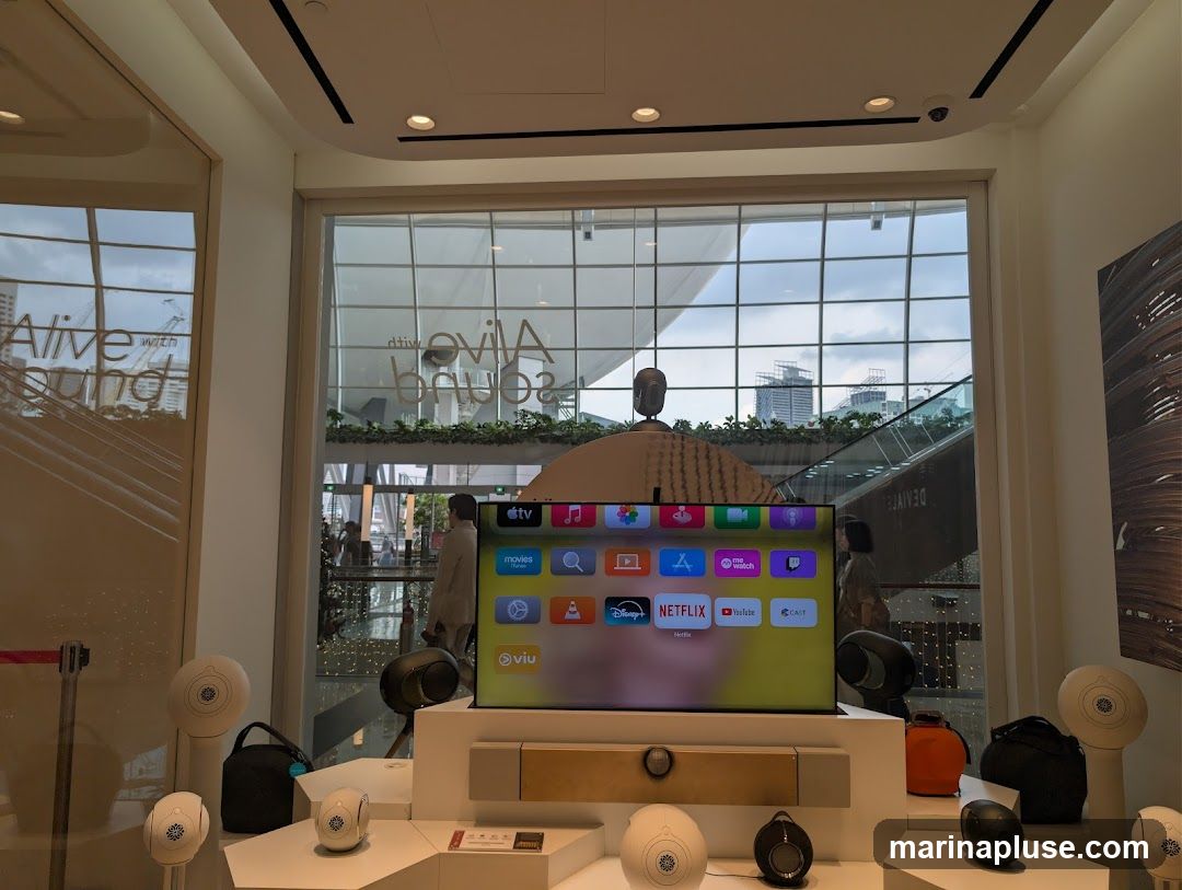 A large television screen displays the Apple TV interface with various app icons, including Apple TV+, Movies, TV Shows, Netflix, YouTube, and Disney+. The screen is part of a retail display featuring several white and black speakers of different sizes arranged around it. In the background, through a large window, a cityscape and an escalator are visible. Text on the window reads "Alive with sound" reflected from the interior.
