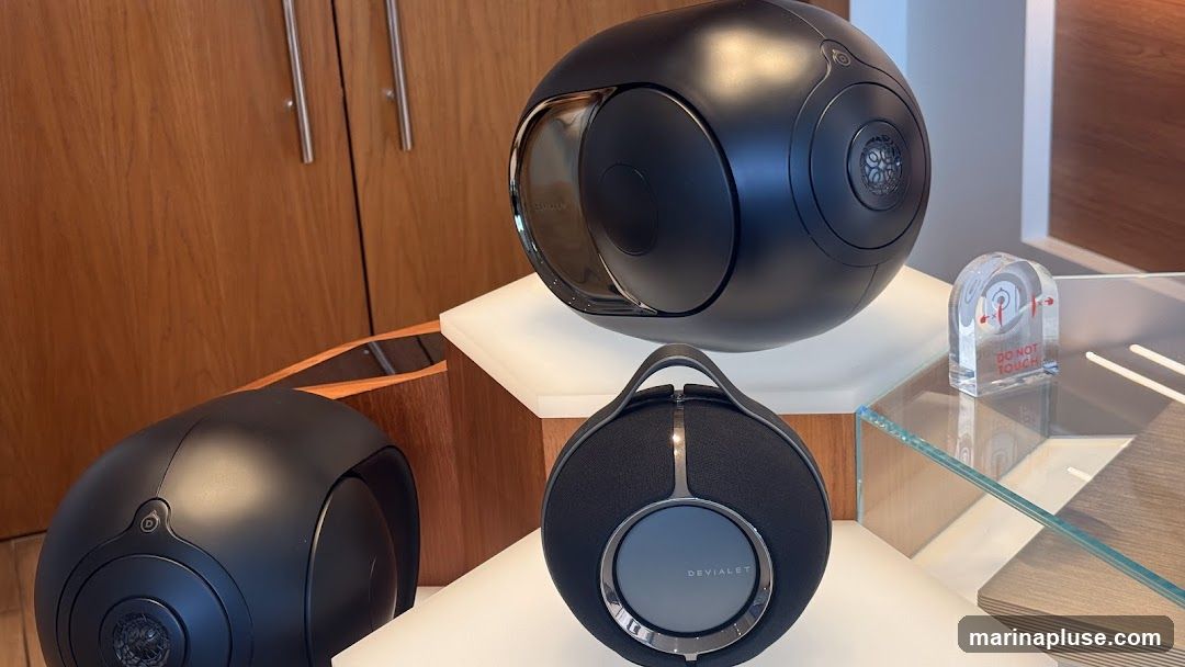 Three black Devialet speakers are displayed on white platforms and glass shelves. One large, spherical speaker is at the top, with two smaller, round speakers below it. A clear acrylic block with red text that reads "DO NOT TOUCH" is placed next to the top speaker. The background features wooden cabinetry.
