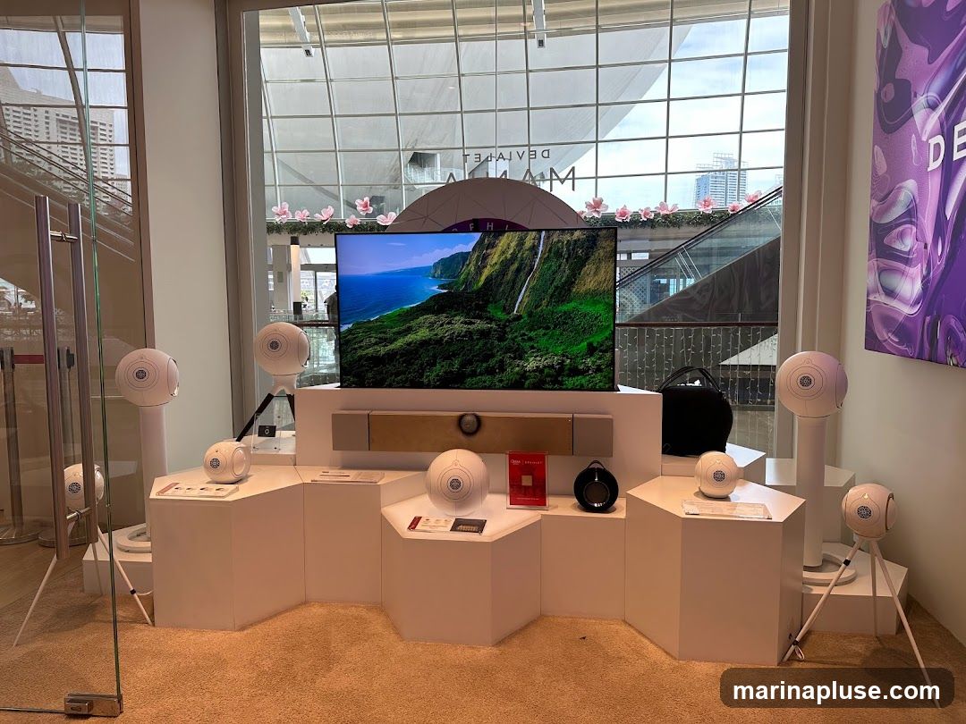 A display of white, spherical speakers and a soundbar in front of a large television showing a scenic landscape. The display is set up on white hexagonal platforms. A black bag is visible behind the television. A purple abstract art poster is on the right wall. The background shows a modern interior with large windows and escalators.