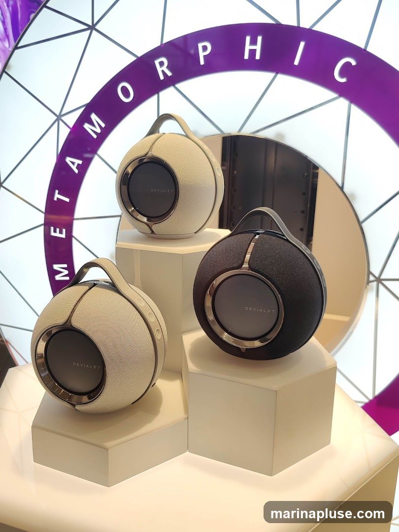 Three spherical Devialet speakers are displayed on white geometric stands. Two speakers are light gray and one is black. A large purple arch with white lettering that reads "METAMORPHIC" is behind the speakers.