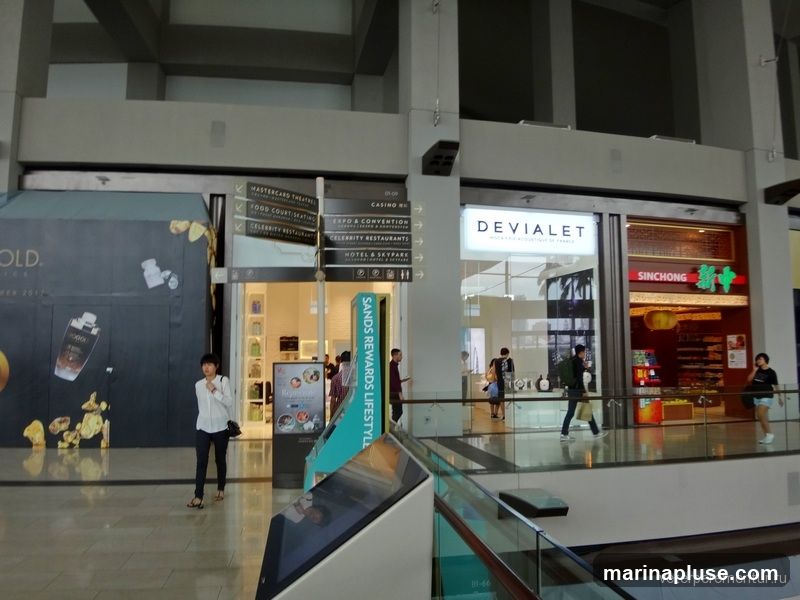 An interior view of a modern shopping mall with various storefronts and signage. On the left, a large advertisement for "GOLD." is visible. To the right, there are signs pointing to "MASTERCARD THEATRES," "FOOD COURT SEATING," "CASINO," "EXPO & CONVENTION," "CELEBRITY RESTAURANTS," and "HOTEL & SKYPARK." Further to the right are storefronts for "DEVIALET" and "SINCHONG." People are seen walking around the mall.