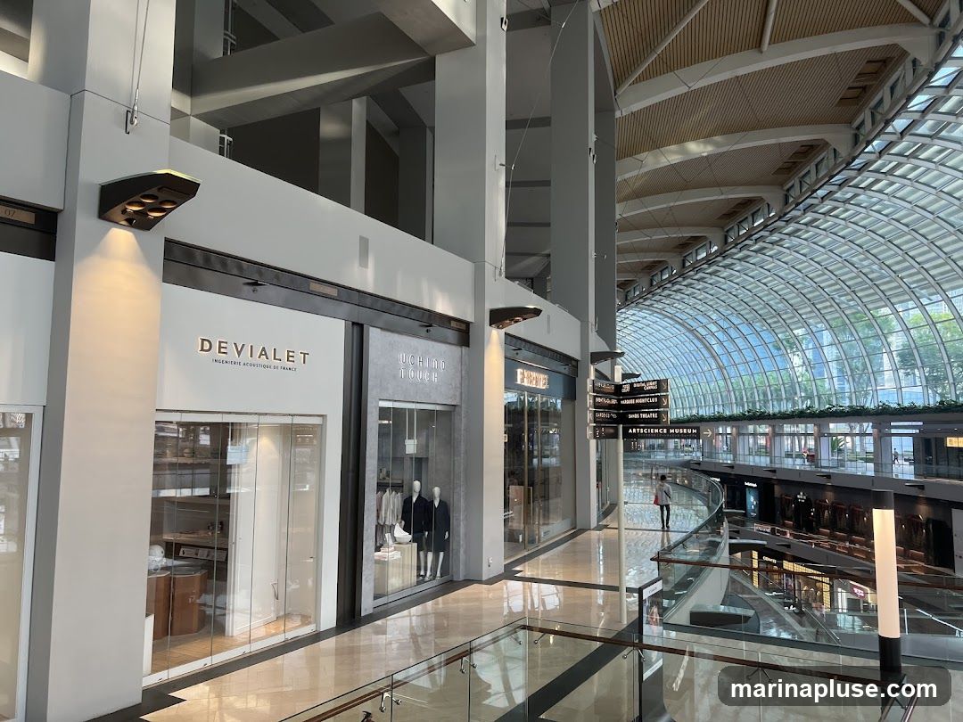 The image shows the interior of a large, modern shopping mall with a distinctive arched glass ceiling. Several storefronts are visible, including one with the name 'DEVIALET' and another with 'UCHINO TOUCH'. Mannequins are displayed in the windows of the 'UCHINO TOUCH' store. Signage indicating directions to various attractions like 'ARTSCIENCE MUSEUM' and 'MARINA BAY SANDS THEATRE' is also present. The mall features polished floors and a multi-level design with glass railings. Natural light streams in through the glass roof, illuminating the spacious interior.
