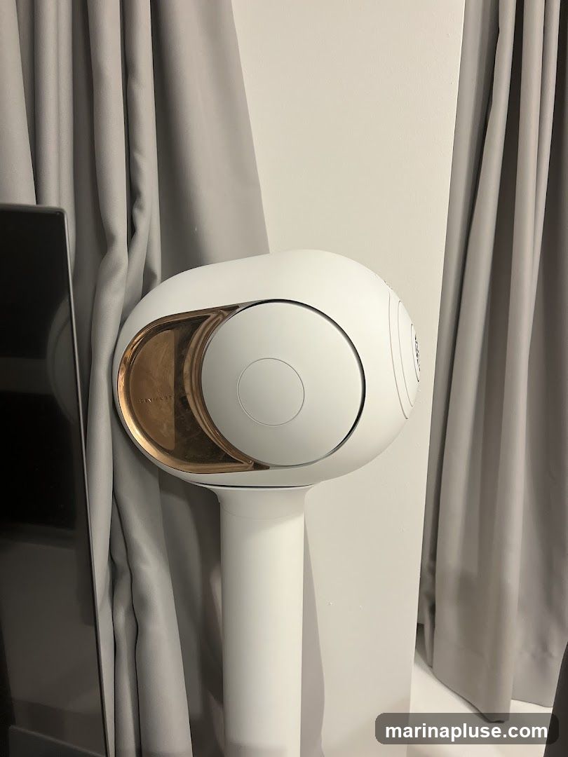A white, spherical speaker with a copper-colored accent is mounted on a white stand. The speaker is positioned next to a gray curtain and a dark, reflective surface, possibly a television screen. The background is a plain white wall.