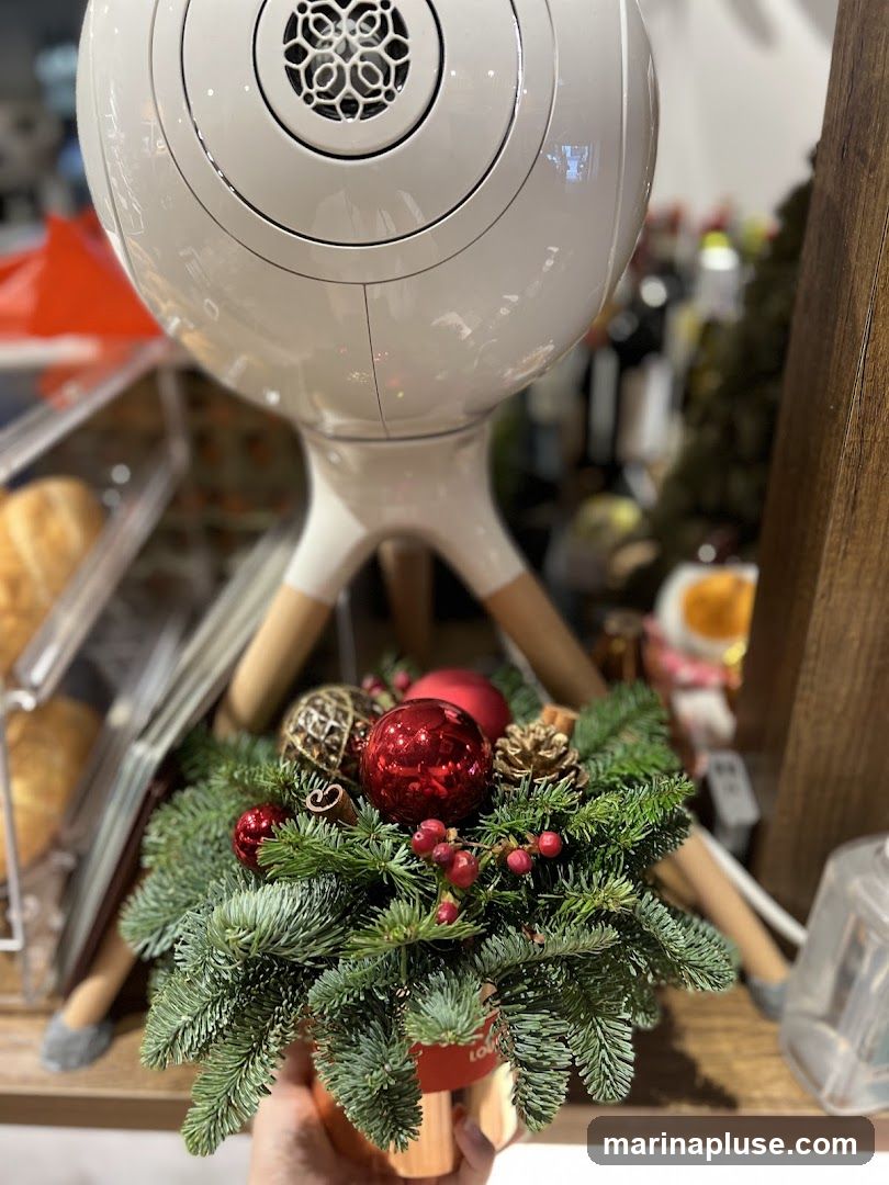 A close-up shot of a Christmas floral arrangement held in a hand, featuring pine branches, red berries, pinecones, and red and gold ornaments. Behind the arrangement, a white, egg-shaped speaker with wooden legs is visible. The background is blurred, showing shelves with items that appear to be bottles and possibly food items, suggesting a retail or display setting. A red tag with some text is attached to the arrangement.