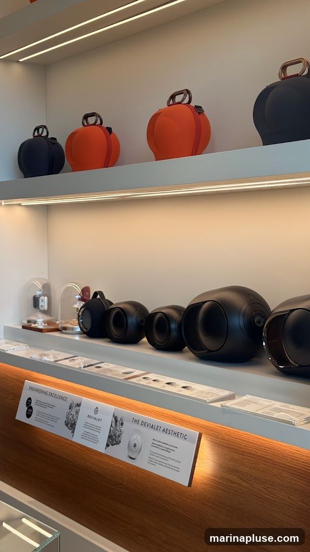 A display shelf featuring various Devialet audio products. On the top shelf, there are several spherical bags in black and orange. Below, on a lower shelf, are several black, oval-shaped speakers. A sign at the bottom reads "ENGINEERING EXCELLENCE" and "THE DEVAILET AESTHETIC" with accompanying text and images.