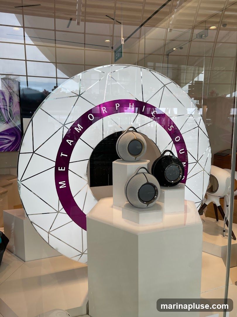A store window display features several spherical speakers on white pedestals. Behind the speakers is a large, circular graphic with geometric patterns and the text "METAMORPHIC SOUND" in purple lettering. The display is viewed through a glass window, with reflections of the interior and exterior visible.