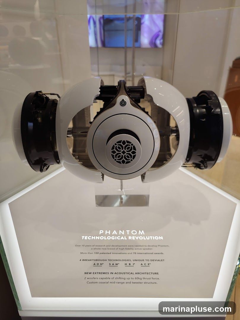 A cutaway view of a white and black Devialet Phantom speaker is displayed in a glass case. Text below the speaker describes its technological revolution, including 4 breakthrough technologies: ADH, SAM, HBI, and ACE. It also highlights new extremes in acoustical architecture with 2 woofers capable of shifting up to 60kg thrust force and a custom coaxial mid-range and tweeter structure.