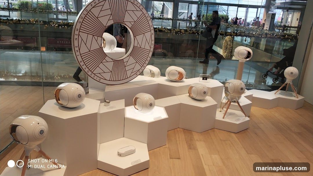 A display of white and gold Devialet Phantom speakers on white hexagonal pedestals. A large circular graphic with the text "DISCOVER THE NEW GOLD PHANTOM" is visible behind the speakers. The display is set up in a modern interior, possibly a shopping mall, with reflections of people and storefronts visible through a glass partition.