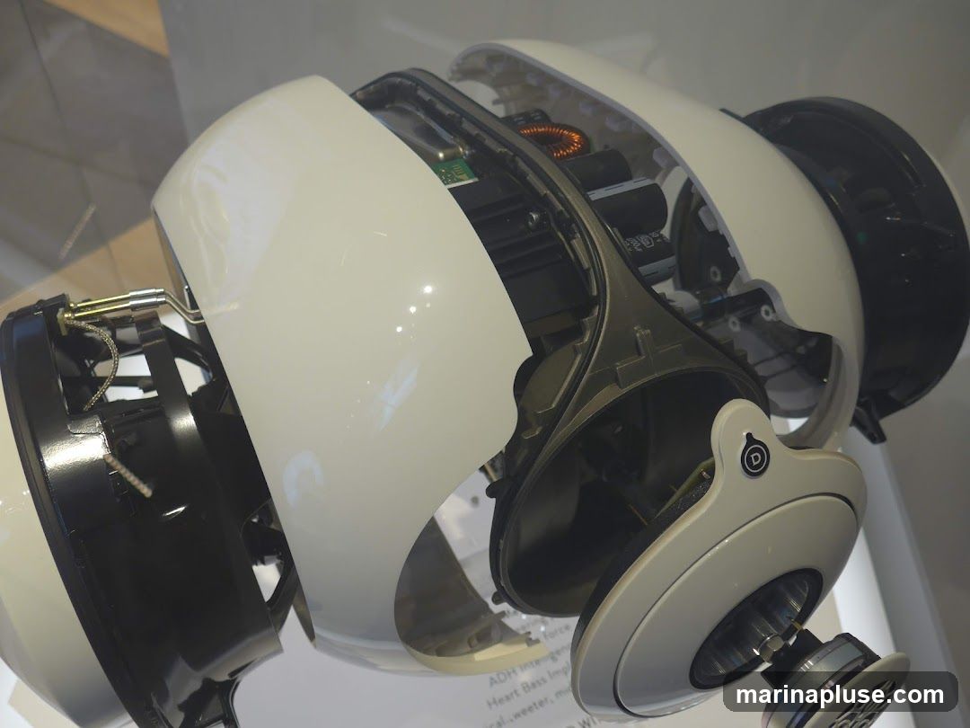 A close-up, detailed view of a deconstructed speaker, showcasing its internal components. The speaker's outer shell is white and glossy, revealing intricate wiring, circuit boards, and a copper coil. The internal structure appears to be made of dark gray and black plastic and metal. A small circular logo with a stylized 'D' is visible on one of the white outer panels. Text is partially visible at the bottom of the image, suggesting technical specifications or branding.