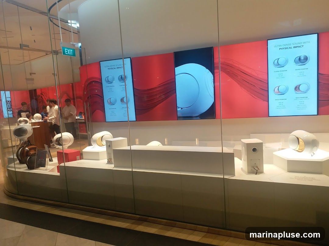 A display of modern speakers in a store. The speakers are white and gold, with some in brown. There are screens in the background showing images of the speakers and text about their sound quality. An "EXIT" sign is visible on the left.