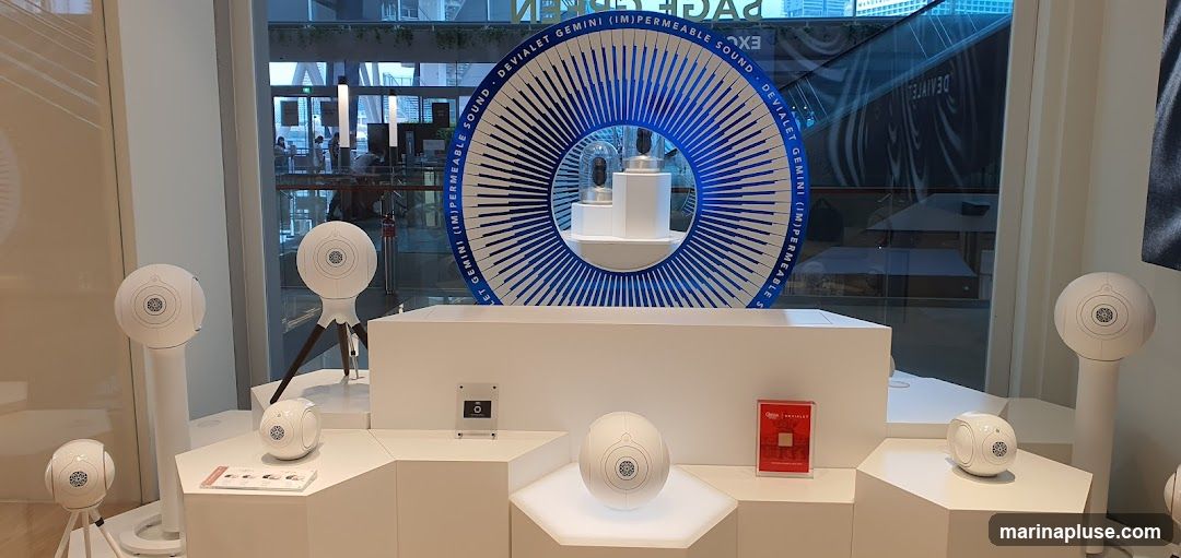 A retail display featuring multiple white spherical speakers of various sizes, some on stands and some on tripods. In the background, a large circular display with blue and white radial lines is visible, with the text "DEVIALET GEMINI (IM)PERMEABLE SOUND" around its circumference. Inside the circle, two smaller spherical objects are displayed. A small red box with a gold component is also on display. The scene appears to be inside a store, with reflections of the outside visible in the windows.