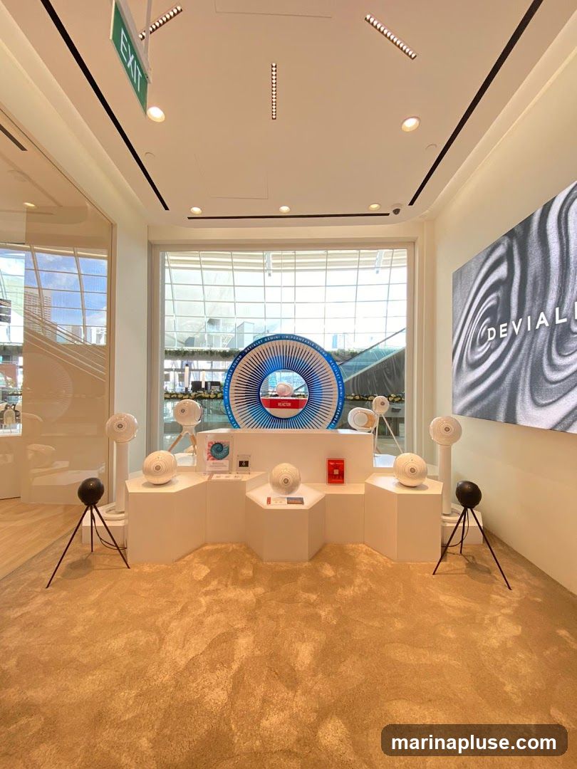 A retail display featuring various spherical speakers and a large circular graphic with the brand name "DEVIALET" visible on a screen. The display is set up on a white tiered platform with a plush beige carpet. An "EXIT" sign is visible on the ceiling.