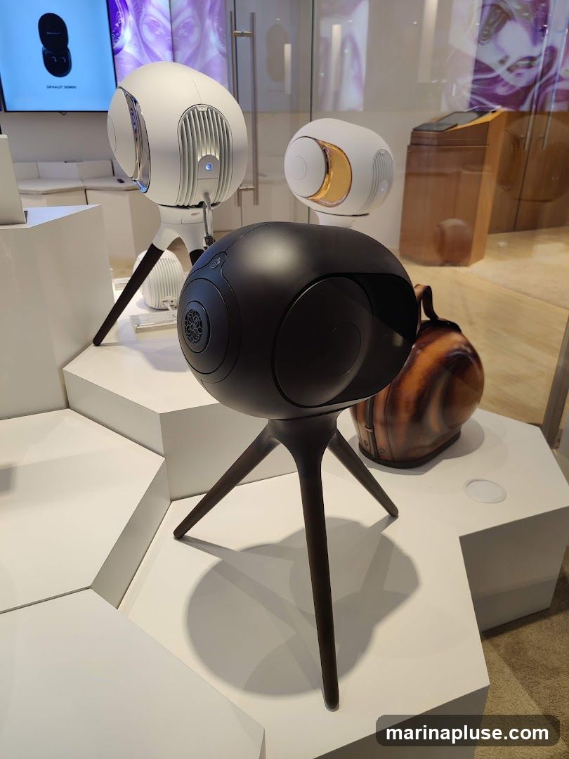 A display of sleek, modern speakers, some white and some black, are showcased on white hexagonal pedestals. In the background, a screen shows an advertisement for 'DEVIALET SONOS'. A decorative, brown, helmet-like object is also visible.