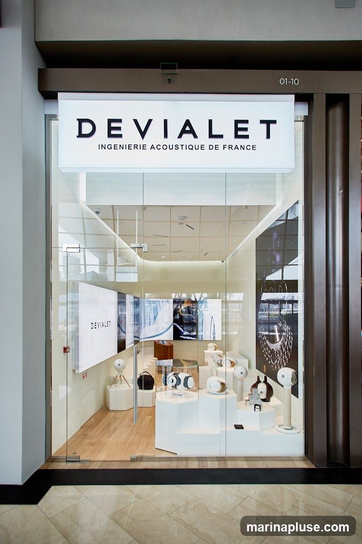 A well-lit storefront with a large glass facade. Above the entrance, a prominent illuminated sign displays "DEVIALET" in large black letters, with smaller text below reading "INGÉNIERIE ACOUSTIQUE DE FRANCE". Inside, the store is modern and minimalist, featuring white display stands showcasing various audio equipment, including speakers and headphones. The interior is visible through the glass, revealing a clean and organized retail space.