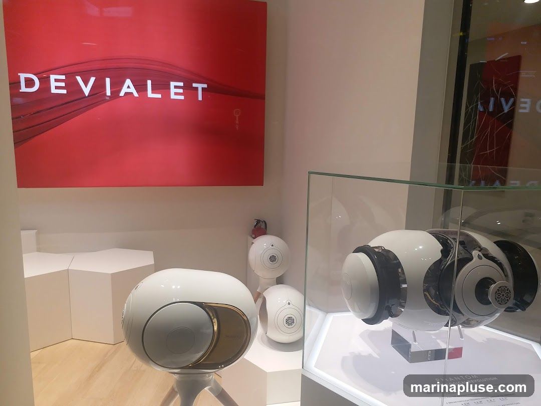 A display window featuring Devialet brand speakers. A large red sign with the Devialet logo and name is prominently displayed. Several white, egg-shaped speakers are arranged on pedestals, with one larger speaker showcased in a glass display case. Text below the larger speaker reads "PHANTOM TECHNOLOGICAL REVOLUTION".