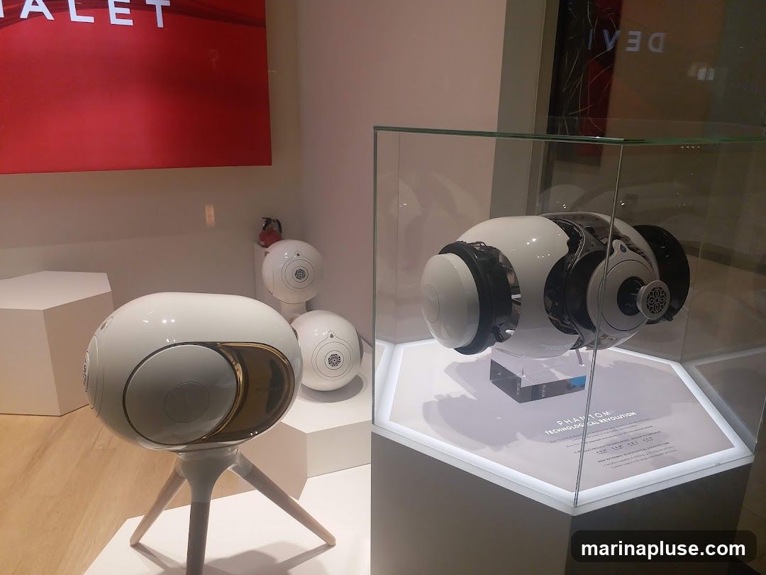 A display of white, spherical Devialet speakers, one on a tripod stand in the foreground, and two smaller ones in the background. A larger, more complex speaker unit is showcased in a glass display case on the right. A red sign with white text is visible on the upper left.