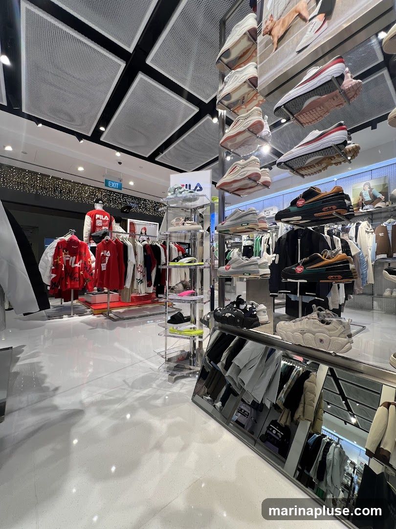 The interior of a Fila store displays various athletic shoes and apparel. Shoes are showcased on tiered shelves, with some displayed on the walls. Clothing racks hold jackets, hoodies, and t-shirts, predominantly in red and white. A prominent Fila logo is visible on a display stand. The store features a clean, modern aesthetic with bright lighting and a reflective floor. An "EXIT" sign is visible in the background.