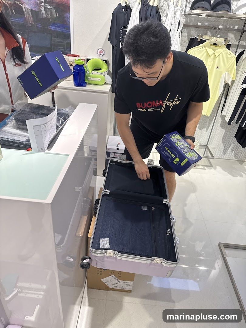 A man in a black t-shirt with a graphic print is examining an open suitcase. He is holding a blue box with the brand name "FUSION" visible. Another person, partially visible on the left, is also holding a similar blue box. The background shows racks of clothing, including t-shirts and hats, with the Fila logo visible on some items. A blue water bottle and a green neck pillow are on a counter. The overall scene appears to be inside a retail store.