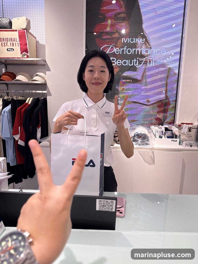 A Fila store employee stands behind a counter, holding a Fila shopping bag and making a peace sign with her fingers. She is wearing a white polo shirt with a Fila logo. Behind her, shelves display Fila merchandise including bags, hats, and clothing. A large digital screen displays an advertisement with the text "Make Performance Beautiful".