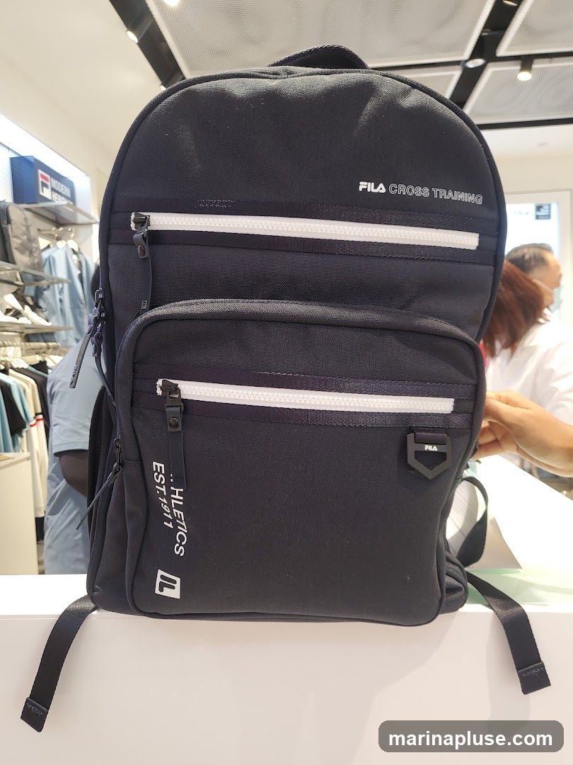 A black Fila backpack with "FILA CROSS TRAINING" and "ATHLETICS EST. 1911" printed on it. The backpack has multiple zippered compartments and is displayed in a store setting with clothing racks and other merchandise visible in the background.