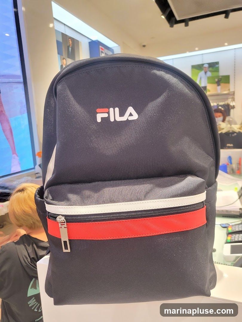 A black Fila backpack with red and white accents is displayed in a store. The Fila logo is visible on the front of the backpack. In the background, there are displays of clothing and promotional images, including a golfer. A person with blonde hair is partially visible to the left.