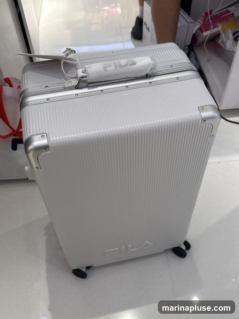 A white Fila suitcase with silver trim and wheels is displayed indoors. The Fila logo is visible on the handle and the front of the suitcase.