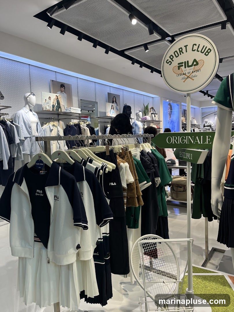 A well-lit interior of a sportswear store, featuring clothing racks filled with Fila branded apparel. Mannequins display outfits, and promotional images of models are visible on the walls. A circular sign with the Fila logo and the text "SPORT CLUB" hangs from the ceiling. Another sign reads "ORIGINALE EST. 1911". A tennis racket display is also present.
