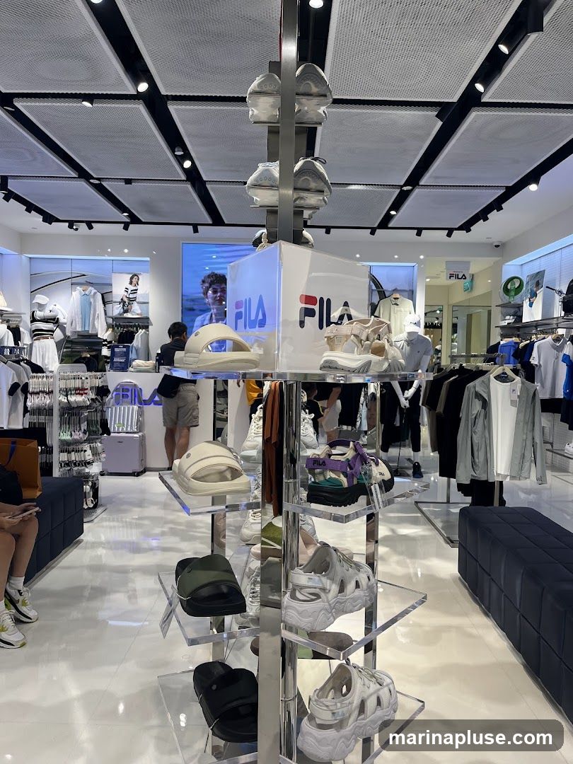 The interior of a Fila store displays various footwear and apparel. Shoes are showcased on tiered shelves, including sandals and chunky sneakers. Mannequins and racks of clothing, such as t-shirts and jackets, are also visible. The store features a modern design with bright lighting and a clean, white floor. A large Fila logo is prominently displayed on a display cube in the center of the image.