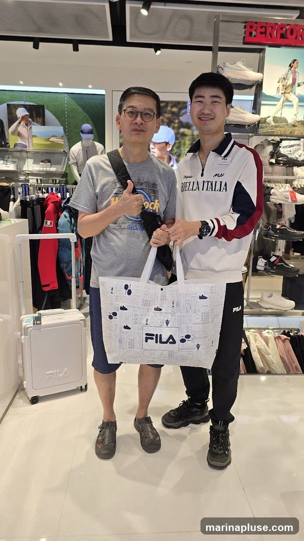 Two men are standing in a Fila store, holding a Fila branded shopping bag. The man on the left is wearing glasses and a grey t-shirt, giving a thumbs up. The man on the right is wearing a Fila branded sweatshirt and black pants. The store interior is visible behind them, with clothing racks and displays of shoes and athletic wear. A white Fila suitcase is on the left.