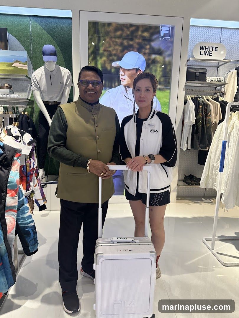 A man and a woman stand in a Fila store, holding a white Fila suitcase. The man is wearing a green vest over a dark shirt and dark pants. The woman is wearing a black and white Fila jacket and shorts. Behind them, there are mannequins dressed in golf attire and racks of clothing. A large screen displays an advertisement for Fila Golf.