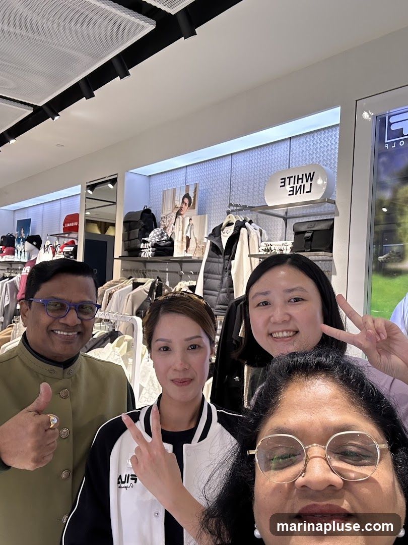 Four people are posing for a selfie in a clothing store. The store appears to be a Fila store, with Fila branding visible on clothing racks and a sign. The people are smiling and making peace signs. The store is well-lit and stocked with apparel and accessories.
