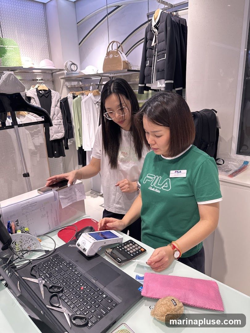 Two women are standing behind a counter in a retail store. One woman, wearing a green Fila t-shirt, is operating a payment terminal. The other woman, wearing glasses and a white t-shirt, is holding a phone. The counter has a laptop, calculator, scissors, and various papers. In the background, clothing and accessories are displayed on racks and shelves.