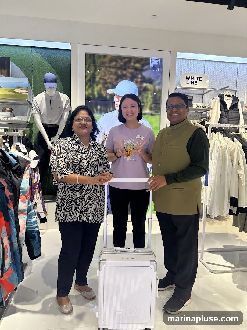 Three people, two women and one man, are standing in a store with a white Fila suitcase. The woman on the left is wearing a black and white patterned shirt and dark pants. The woman in the middle is wearing a light purple t-shirt and dark pants, and is making a peace sign with her fingers. The man on the right is wearing a green vest over a dark shirt and dark pants. They are all smiling. The background shows clothing racks with Fila branded apparel and a large advertisement featuring a golfer.