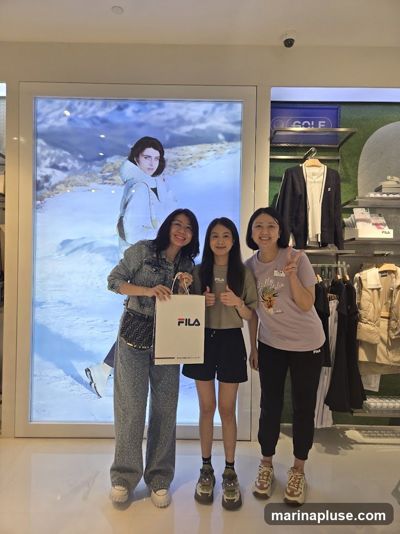 Three people are posing for a photo in a Fila store. The person on the left is wearing a denim outfit and holding a Fila shopping bag. The person in the middle is wearing a grey t-shirt and black shorts. The person on the right is wearing a purple t-shirt and black pants. Behind them is a large display with a model wearing a winter jacket, and racks of clothing.