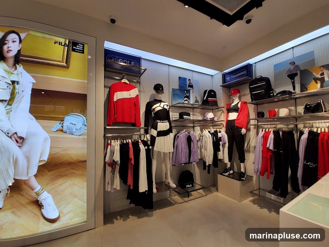 The interior of a Fila store displays athletic apparel on racks and mannequins. A large digital advertisement featuring a model in Fila clothing is visible on the left. Shelves hold backpacks, hats, and accessories. The store is well-lit and organized, showcasing various Fila branded items for golf and training.