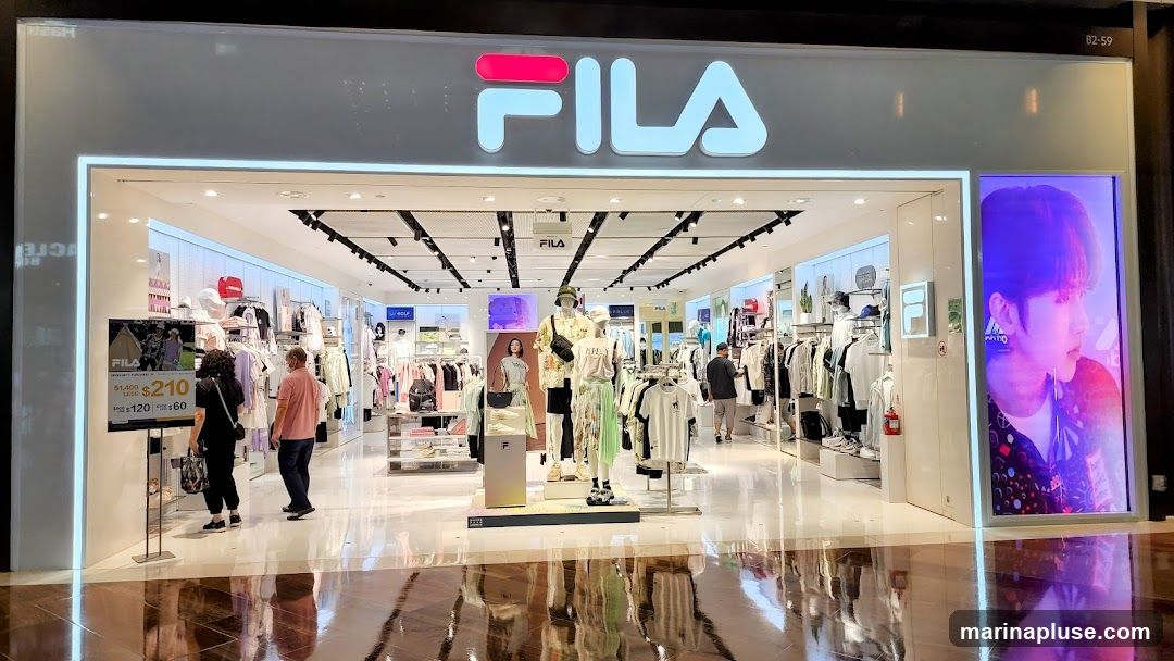 The exterior of a Fila store with the Fila logo prominently displayed above the entrance. The store is well-lit and showcases various apparel and accessories through its large display windows and open entrance. Inside, mannequins are dressed in Fila clothing, and racks of merchandise are visible. A digital display on the right features a close-up of a person's face with vibrant lighting. A promotional sign is placed near the entrance.