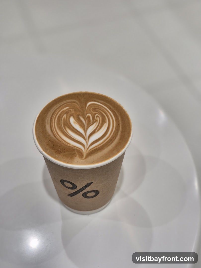 A close-up shot of a latte in a brown paper cup with the percentage logo. The latte has intricate foam art resembling a heart. The cup is placed on a white surface.