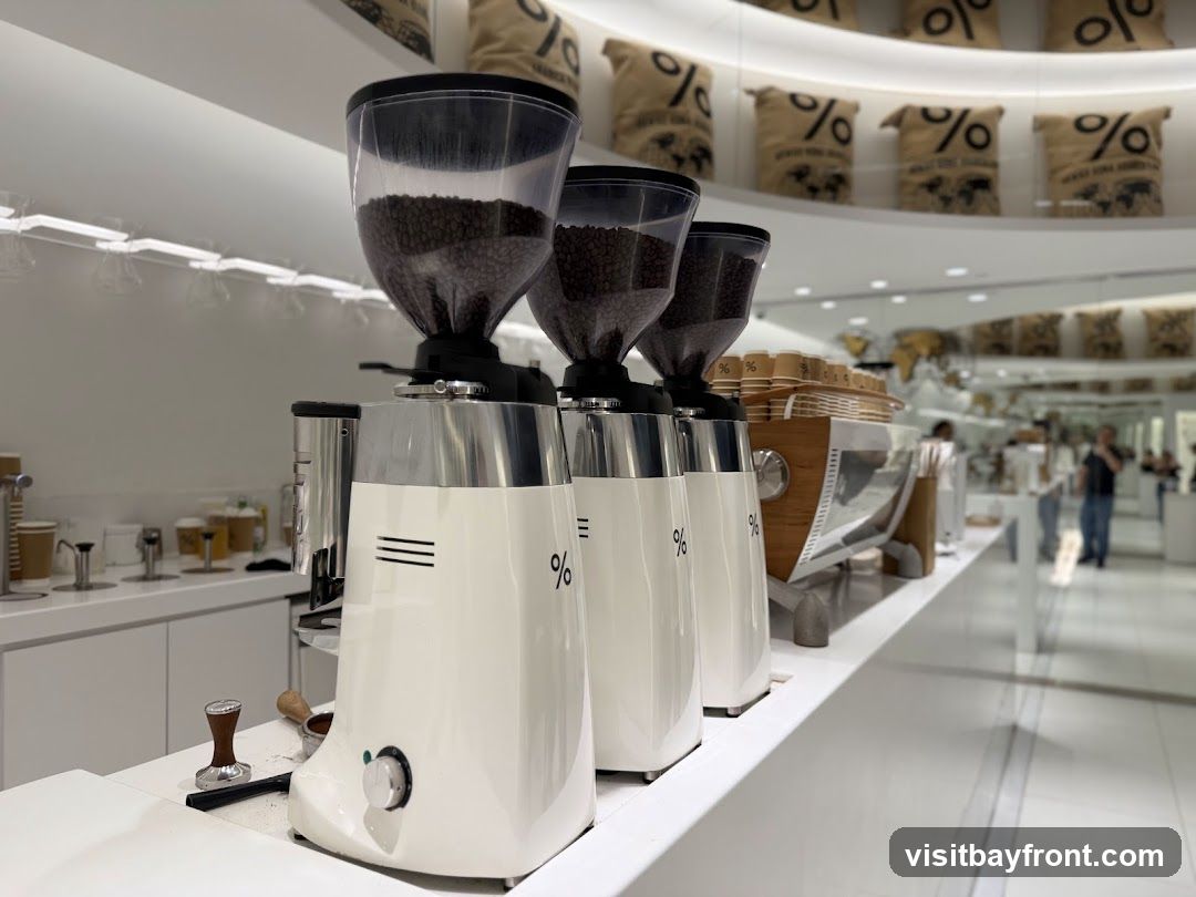 A close-up view of three white coffee grinders with clear hoppers filled with coffee beans, lined up on a white counter. Each grinder has a distinctive percentage logo. Behind them, a wooden-accented espresso machine is visible, along with stacks of paper cups. In the background, shelves display burlap sacks with similar percentage logos, and several people are seen in the distance, suggesting the interior of a coffee shop.