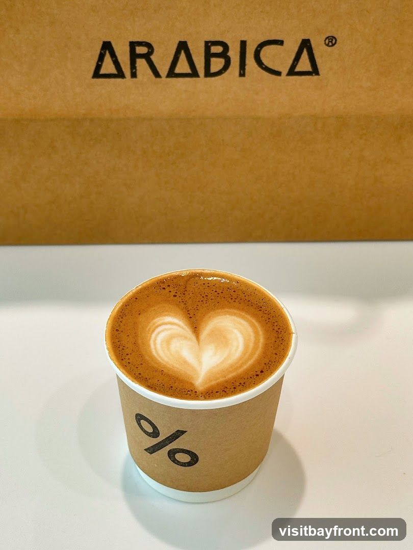 A cup of coffee with latte art in the shape of a heart sits on a white surface. Behind the cup is a brown paper bag with the word "ARABICA" printed in black. The cup itself has a logo that appears to be a percentage sign.