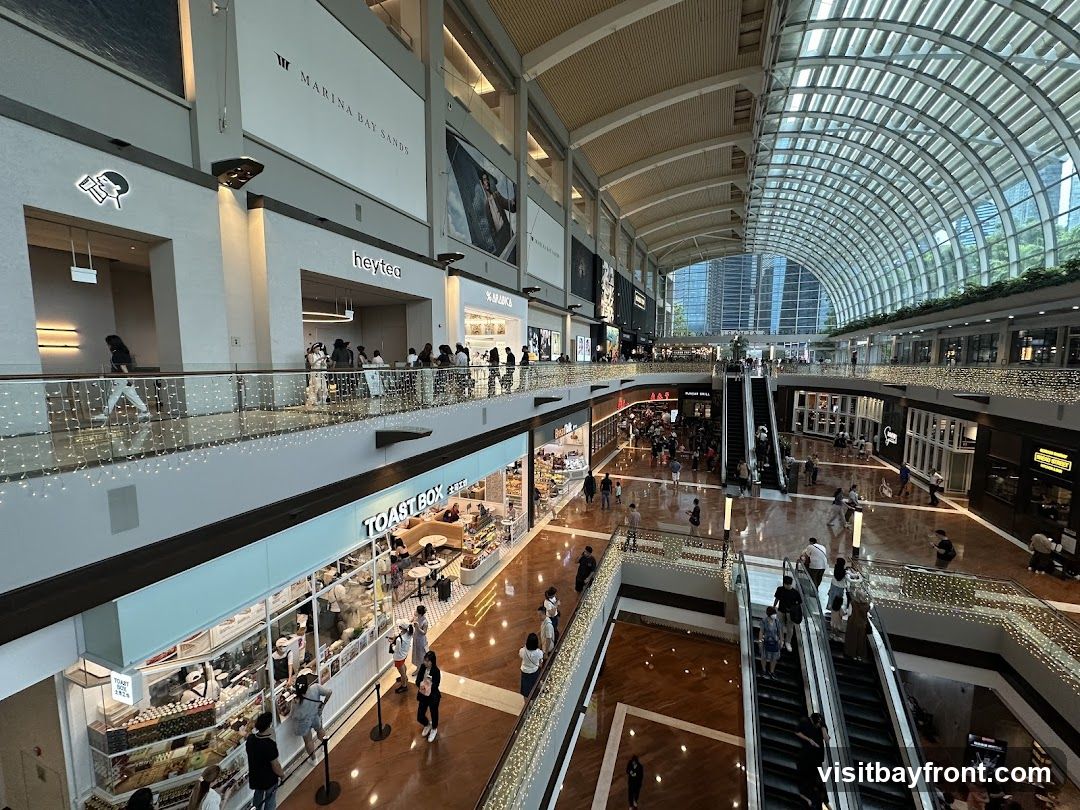 A wide-angle view of the interior of a multi-level shopping mall, featuring a glass-domed ceiling, escalators, and various shops. People are seen walking and shopping on different levels. The mall is decorated with string lights. Prominent signage includes 'MARINA BAY SANDS', 'heytea', and 'TOAST BOX'.