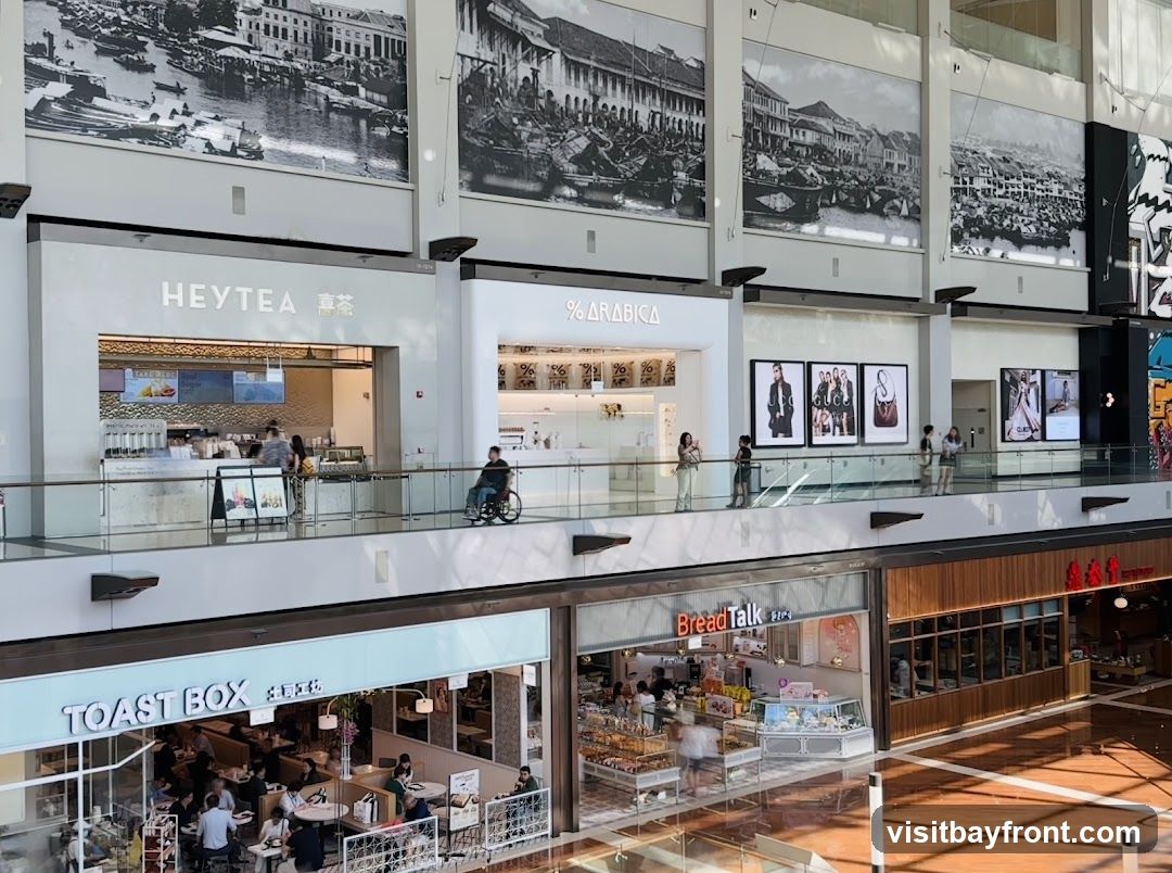 A view of a multi-level shopping mall interior. On the upper level, storefronts for HEYTEA and % ARABICA are visible, with large black and white historical murals above them. Further along, framed fashion advertisements are displayed. On the lower level, the storefronts for TOAST BOX and BreadTalk are prominent, showcasing their food offerings and seating areas. People are seen walking and dining in the mall.