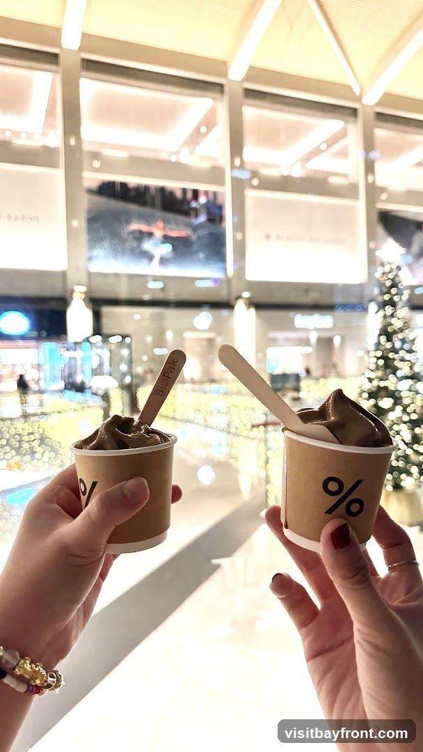 Two hands hold up cups of chocolate soft serve ice cream. The cups are brown and have a black percentage sign logo. Wooden spoons with the logo "BioPak" are in each cup. The background appears to be a mall or shopping center with Christmas decorations, including a decorated tree and string lights.